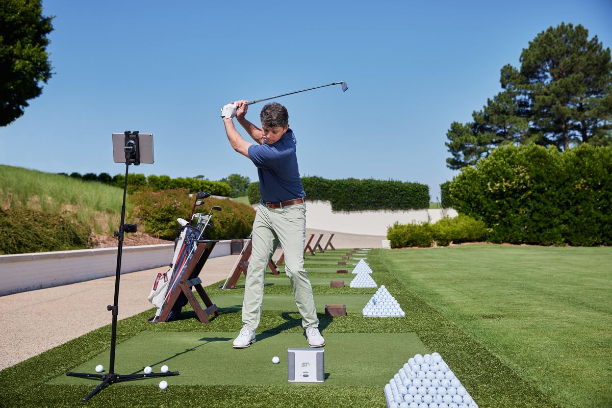 Golfer beim Abschlag auf Driving Range mit Golfbällen, Golftasche und Golf Simulator Outdoor