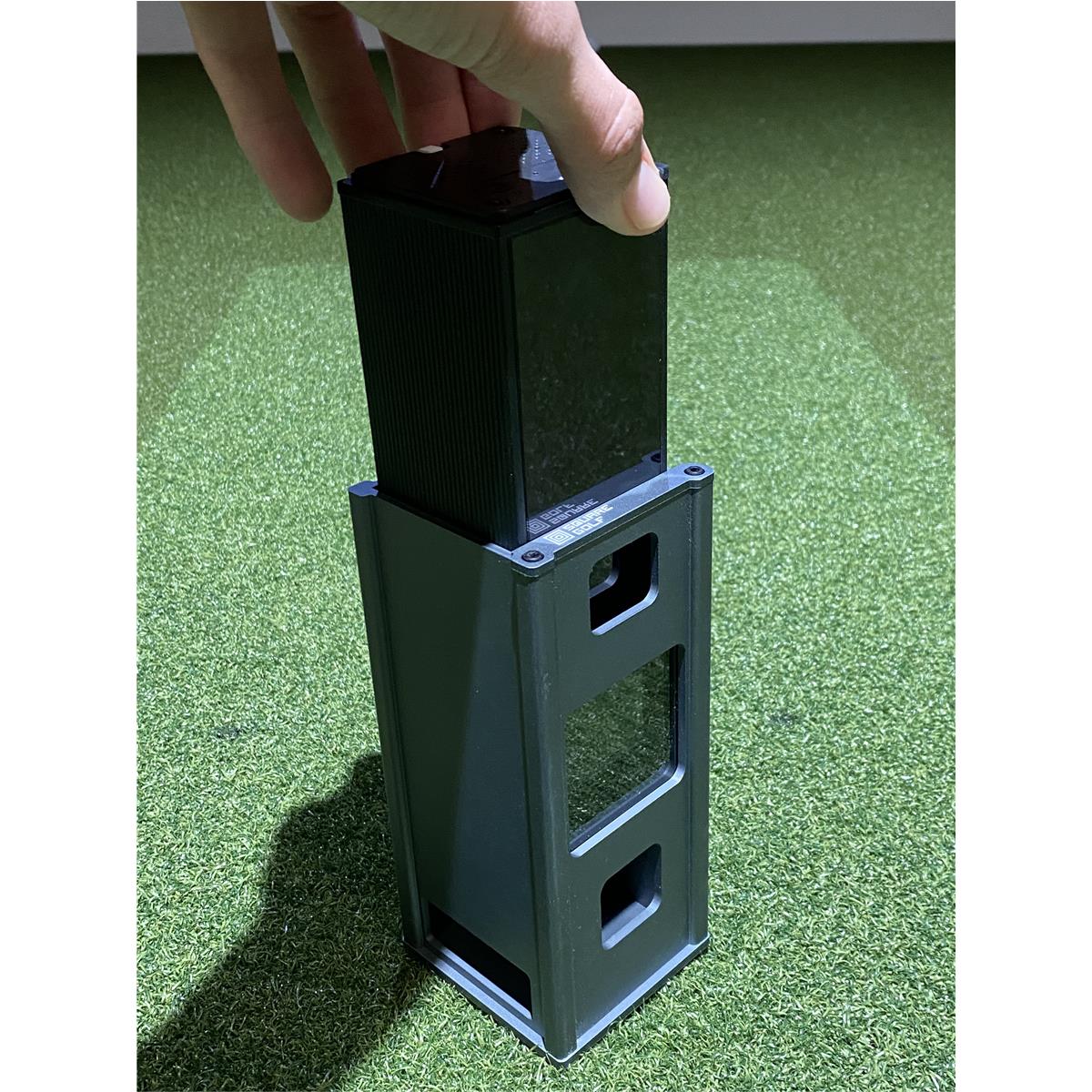 Hand hält schwarzen Akku für Golf Simulator auf grünem Indoor Kunstrasen